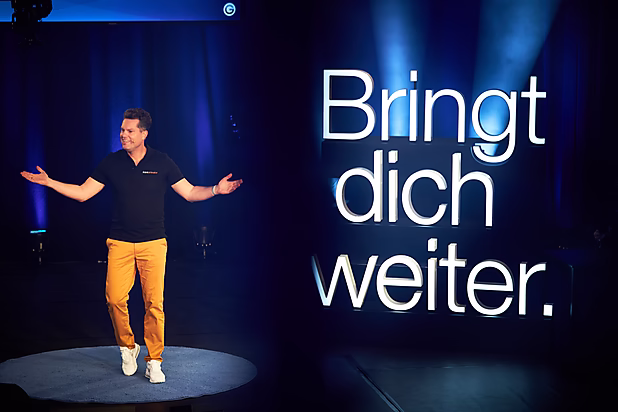 Ein Mann in schwarzem T-Shirt und orangefarbenen Hosen steht auf einer Bühne und präsentiert den Slogan: „Bringt dich weiter.“