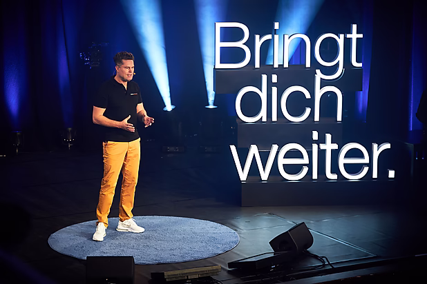 Ein Redner steht auf einer Bühne vor großen, hellen Buchstaben mit dem Text "Bringt dich weiter." Er vermittelt eine motivierende Botschaft.