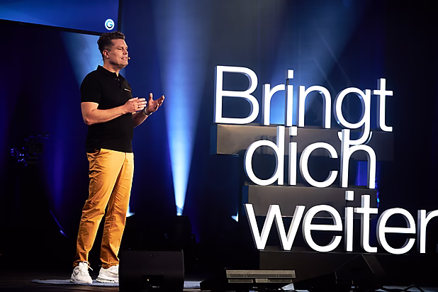 Ein Mann spricht auf einer Bühne, während hinter ihm in großen Buchstaben steht: "Bringt dich weiter".