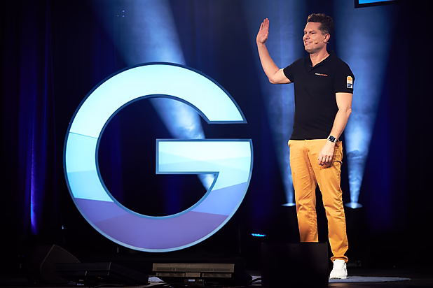 Ein Mann in schwarzer T-Shirt und orangefarbener Hose steht auf einer Bühne und winkt, im Hintergrund ein großes, beleuchtetes "G".