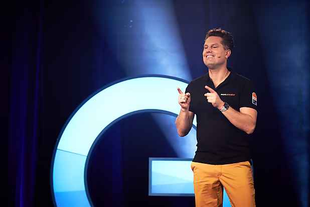 Ein lächelnder Mann in schwarzem T-Shirt und orangenen Shorts hält einen Vortrag auf der Bühne vor einem großen, beleuchteten Logo.