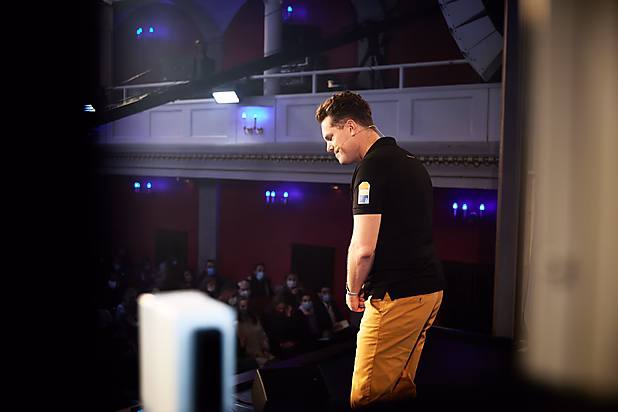 Ein Mann in schwarzem Shirt und gelben Hosen steht auf einer Bühne und schaut nach unten. Publikum im Hintergrund.