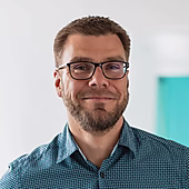 Ein Mann mit Brille und Bart lächelt in einem karierten Hemd. Im Hintergrund ist eine unscharfe, helle Umgebung zu sehen.