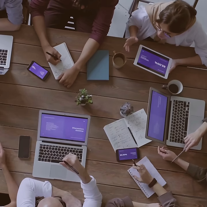 Ein Team sitzt um einen Holztisch und arbeitet gemeinsam an einer digitalen Marketing-Strategie. Mehrere Laptops, Tablets und Smartphones zeigen ein einheitliches Präsentationsdesign, während Notizen auf Papier gemacht werden.