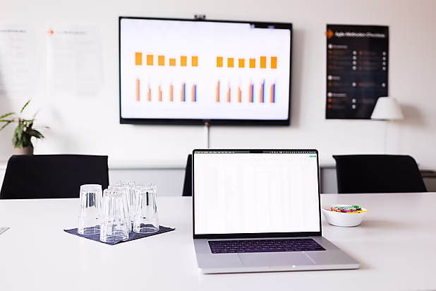 Blick in einen Konferenzraum: AUf dem Tisch steht ein Laptop, auf dem eine Tabelle angezeigt wird, sowie Gläser und ein Schalen voller Süßigkeiten. Im Hintegrurnd werden Diagramme auf einen Bildschirm gezeigt.