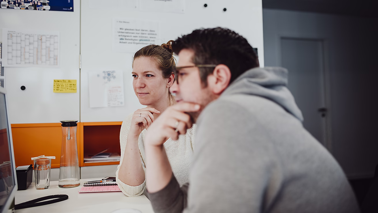 Zwei Personen sitzen an einem Tisch, schauen konzentriert auf einen Computerbildschirm.