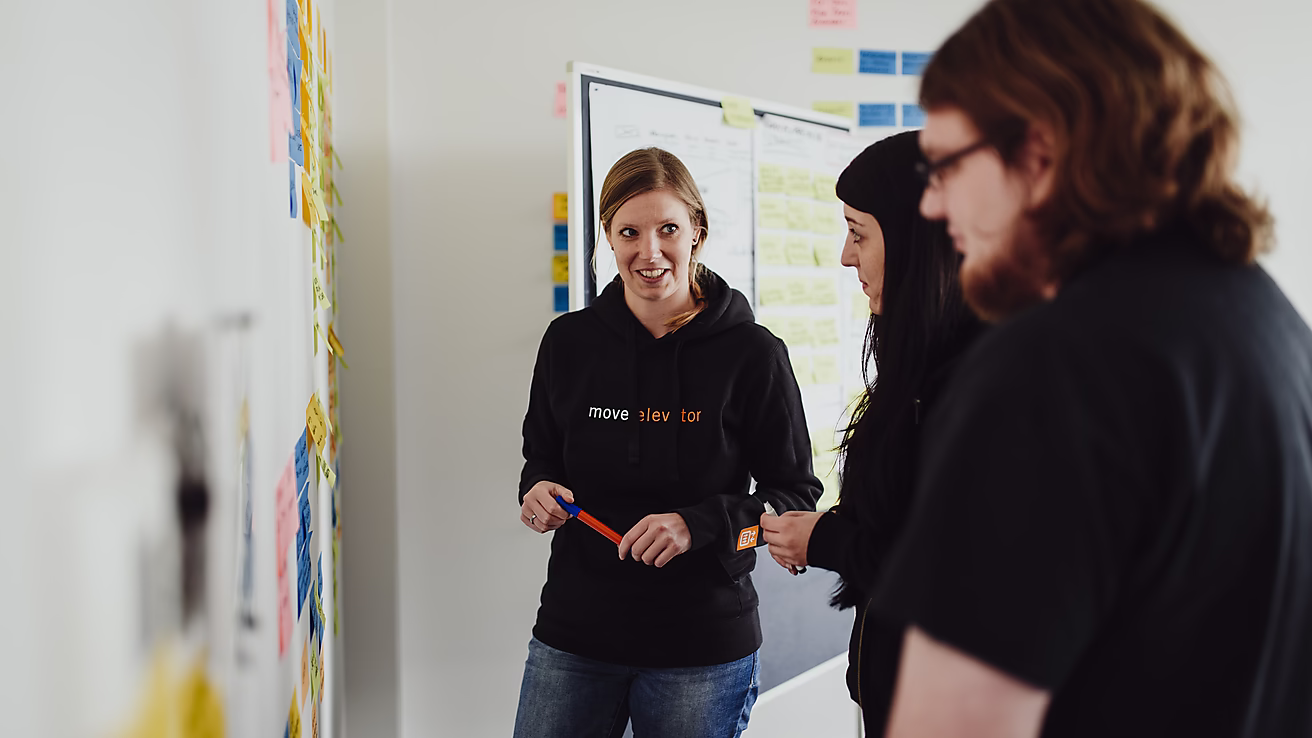 Drei Mitarbeiter brainstormen gemeinsam für ein Projekt. An der Wand kleben viele bunte Haftnotizen mit Ideen und Aufgaben.