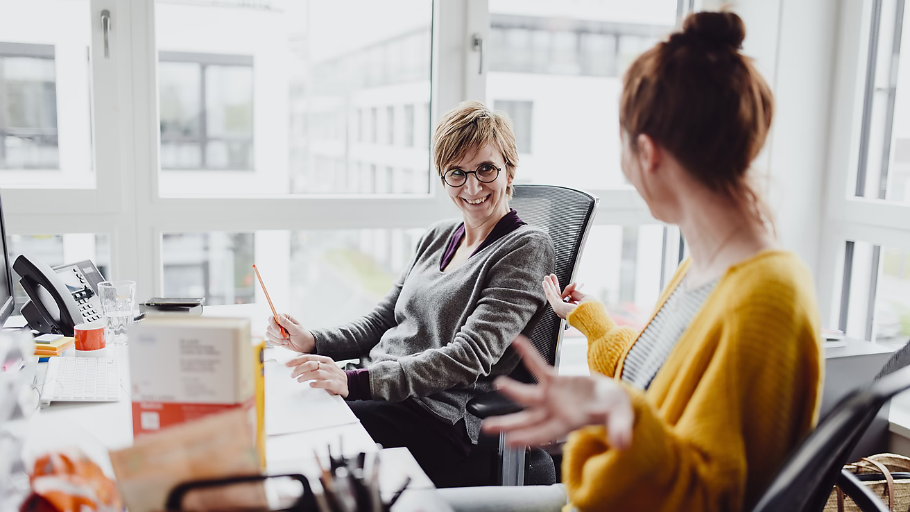 Zwei Frauen sitzen in einem Büro und unterhalten sich freudig. Eine Frau lächelt, die andere gestikuliert mit der Hand.