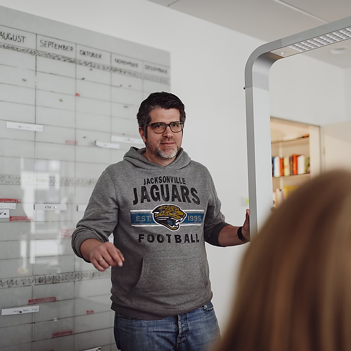 Ein Mann spricht in einem Besprechungsraum vor einem Planungsboard. Vor ihm sitzt eine weitere Person.