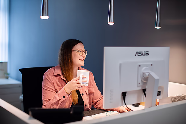 Eine Frau sitzt lächelnd an einem Schreibtisch, hält eine Tasse und schaut auf einen Computerbildschirm in einem modernen Büro.
