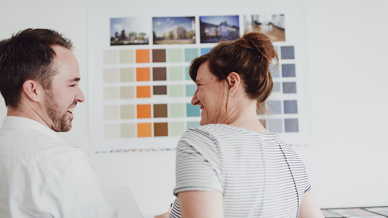 Ein Mann und eine Frau schauen lächelnd auf eine Farbpalette an der Wand, während sie sich unterhalten.