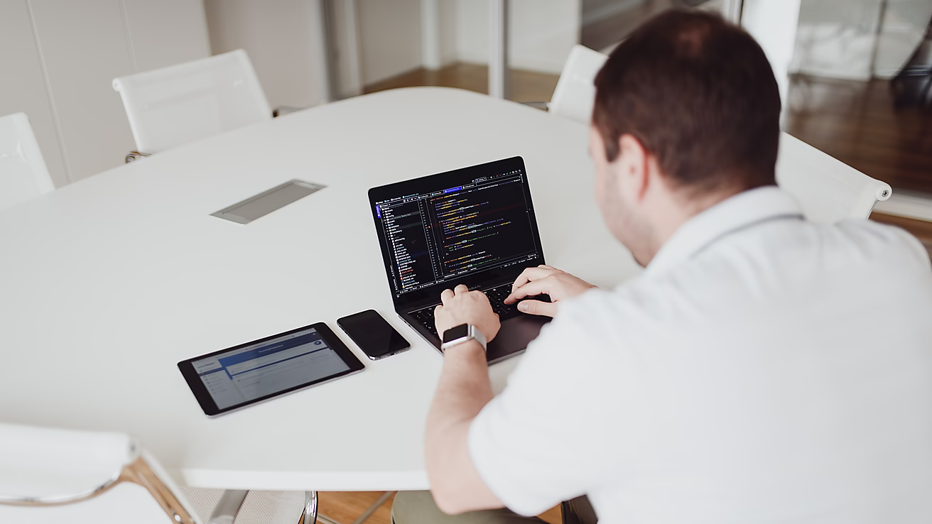 Ein Mann sitzt an einem Tisch und arbeitet auf einem Laptop, während ein Tablet und ein Smartphone daneben liegen.
