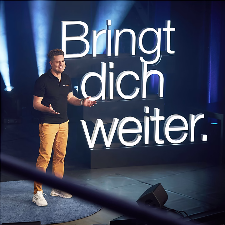 Ein Redner steht auf einer Bühne, hinter ihm leuchtet der Schriftzug "Bringt dich weiter." in großer Schrift.