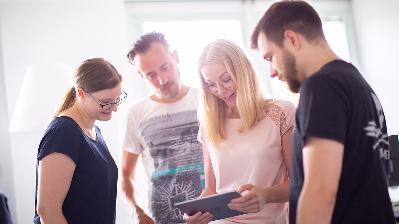 Vier Personen schauen gemeinsam auf ein Tablet und diskutieren interessiert darüber in einem hellen Raum.