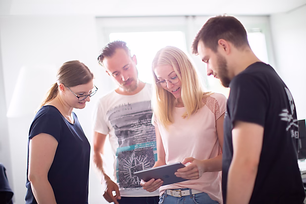 Vier Personen schauen gemeinsam auf ein Tablet und diskutieren interessiert darüber in einem hellen Raum.