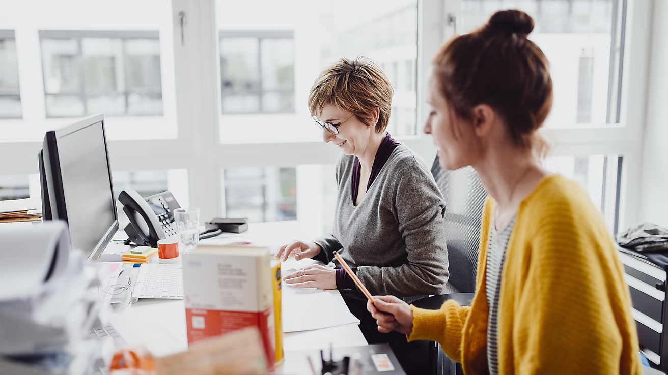 Zwei Frauen sitzen an einem Schreibtisch, arbeiten zusammen und lächeln während ihrer Besprechung in einem hellen Büro.