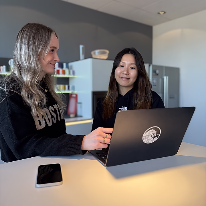 Zwei Frauen sitzen an einem Tisch, eine schaut auf ihren Laptop und erklärt etwas, die andere lächelt. Ein Smartphone liegt auf dem Tisch.