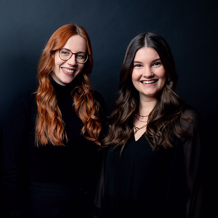 Zwei Frauen stehen vor einem dunklen Hintergrund und lächeln freundlich. Eine hat lange, rote Haare, die andere hat lockeres, braunes Haar.