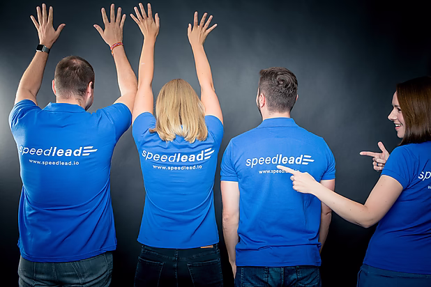 Vier Personen in blauen Poloshirts mit dem Logo "speedlead" zeigen in die Höhe. Eine Person lächelt und deutet mit dem Finger auf ihre Kollegen. Es herrscht eine lockere, zwanglose Atmosphäre.