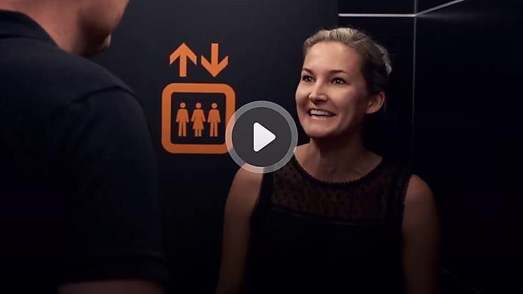 Eine Frau lächelt in einem Aufzug, während sie mit jemandem spricht. Im Hintergrund sind Aufzugs- und WC-Symbole sichtbar.