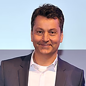 Ein lächelnder Mann in einem dunklen Anzug mit weißem Hemd steht vor einem neutralen Hintergrund.