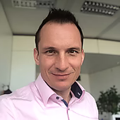 Ein Mann mit kurzem, dunklem Haar trägt ein pinkes Hemd und lächelt in die Kamera, vor einem modernen Büro-Hintergrund.