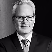 Porträt eines Mannes in formeller Kleidung mit Brille, lächelnd vor einem dunklen Hintergrund.
