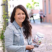 Eine lächelnde Frau hält einen Informationsflyer in einer malerischen Gasse.