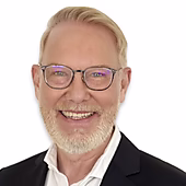 Ein lächelnder Mann mit Bart und Brille trägt einen schwarzen Blazer und ein weißes Hemd vor einem neutralen Hintergrund.