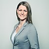 Eine professionelle Frau in einem grauen Blazer lächelt und steht vor einem hellen Hintergrund.