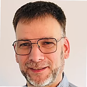 Ein lächelnder Mann mit Brille und gestutztem Bart, vor einem hellen Hintergrund.