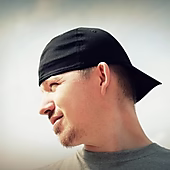 Ein Mann mit einem schwarzen Baseballcap schaut nach rechts und lächelt leicht, im Hintergrund ein heller, wolkiger Himmel.