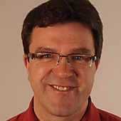 Ein lächelnder Mann mit Brille trägt ein rotes Hemd, vor einem neutralen Hintergrund.