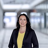 Frau mit langen schwarzen Haaren in Blazer und gelbem Oberteil steht in einem modernen Innenraum mit vielen Fenstern.